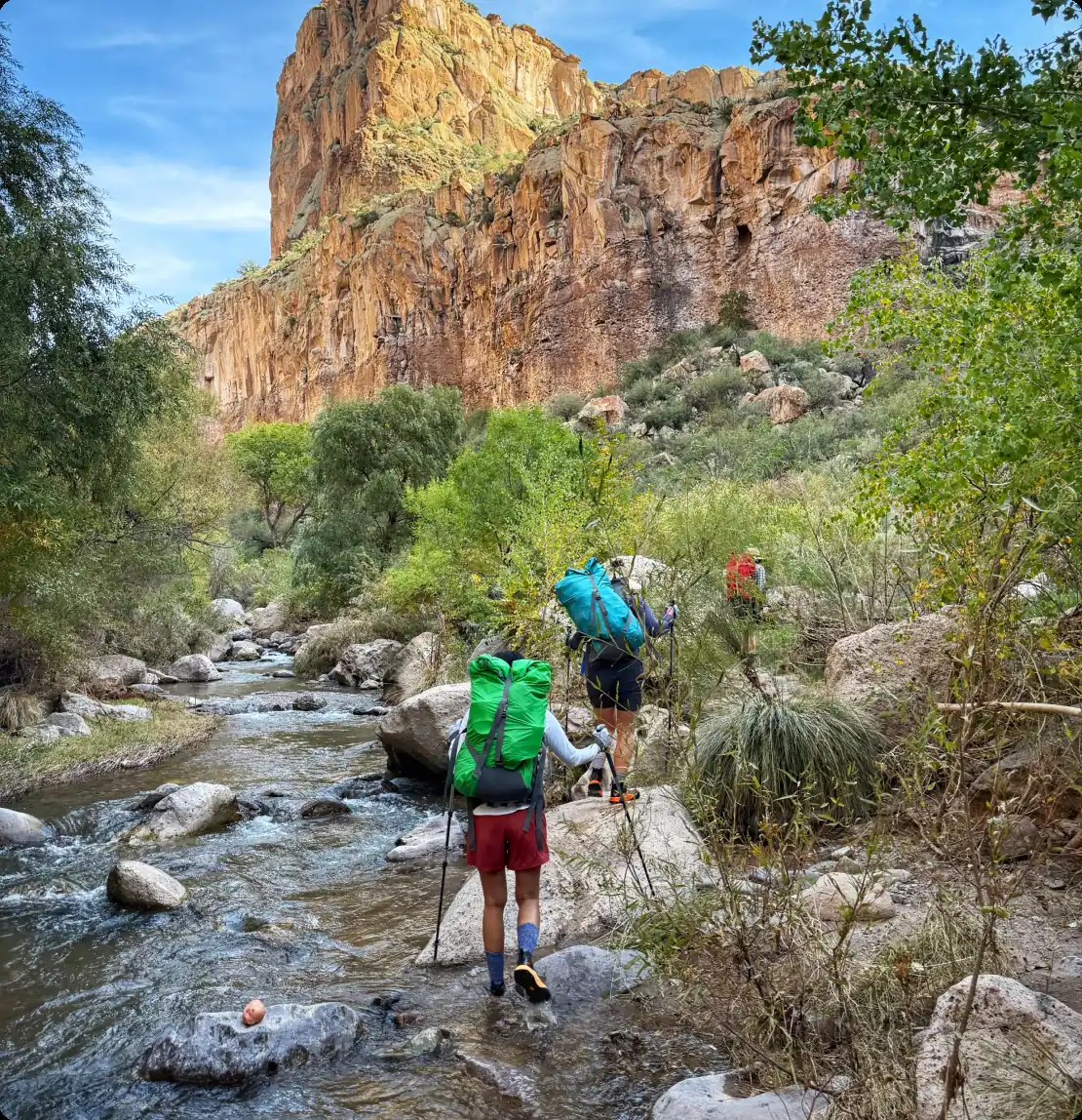Group of backpackers wading Aravaipa Creek below a towering red cliff